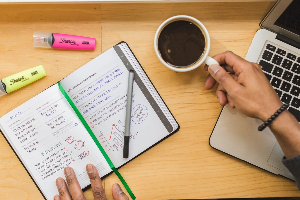Manos escribiendo en un cuaderno junto a un portátil y un café